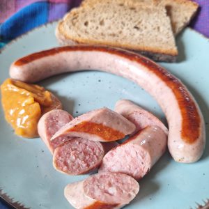 Gegrillte Bratwurst mit Senf und Brotscheiben auf einem Teller vor buntem Tuch
