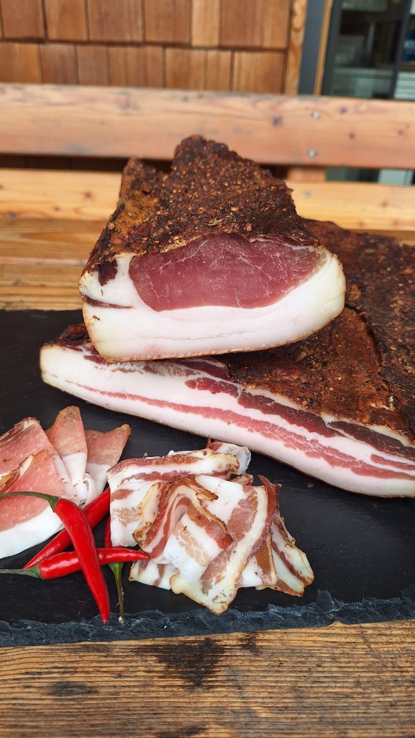 Lungauer Speck auf Schieferplatte mit Pfefferoni und Holzbank im Hintergrund